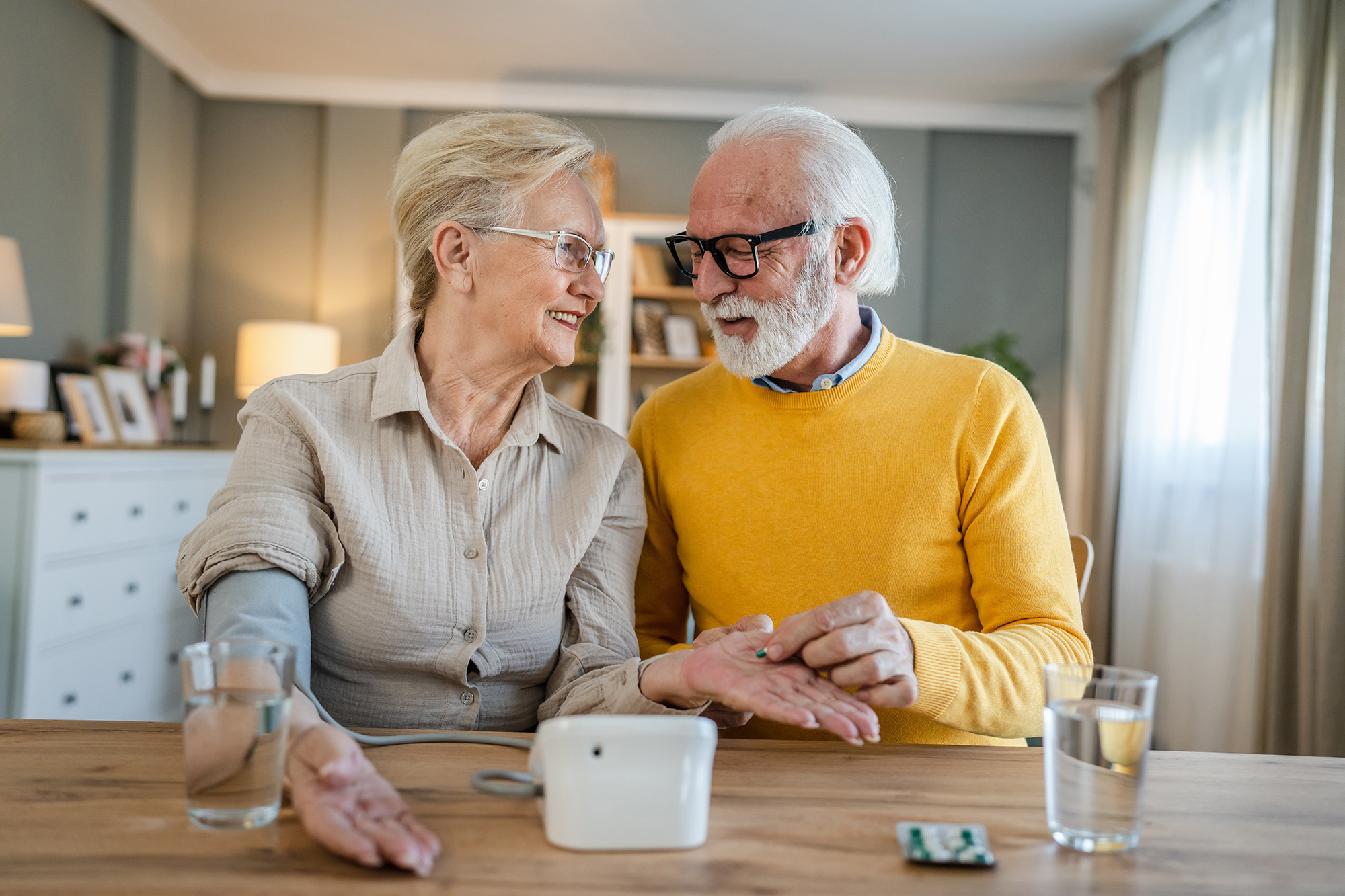 Senior couple woman check measure blood pressure while her husband old caucasian man sit beside at the table at home real people family old people retirement and health concept